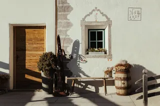 Ostí Vedl sunlit rustic facade with a wooden door, small window adorned with flowers, vintage skis leaning, wooden bench, and barrel planter.